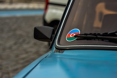 A close-up view of a blue car with a side mirror visible. On the windshield, there's a sticker featuring the flag of Azerbaijan. The background is out of focus, but part of another vehicle can be seen.