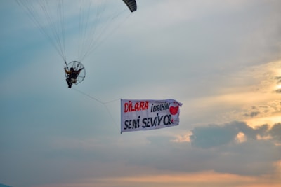 A person is flying a powered paraglider in the sky, holding a large banner. The banner has the names 'Dilara' and 'Ibrahim' along with a heart symbol, conveying a message of love. The sky is partly cloudy with a mix of blue and orange hues from the setting or rising sun.