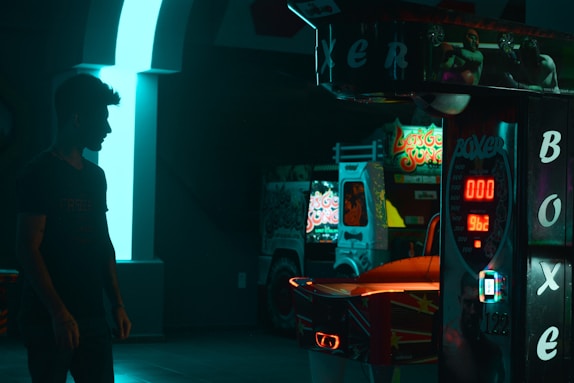 A dark, arcade-like environment with neon lighting highlights. A person is silhouetted in the foreground, facing a punching arcade machine that displays the word 'Boxer' and a digital score. In the background, parts of other arcade machines are visible, adding to the vibrant and lively atmosphere.