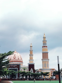 A large mosque complex featuring a grand dome with intricate patterns and two towering minarets. The mosque is surrounded by lush green trees and has a sign reading 'Masjid Agung' at the entrance. The sky is overcast, creating a serene and peaceful atmosphere.