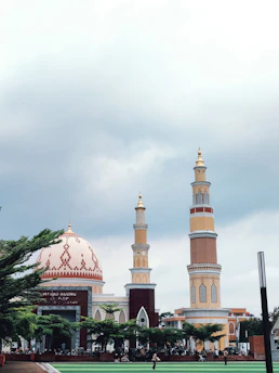A large mosque complex featuring a grand dome with intricate patterns and two towering minarets. The mosque is surrounded by lush green trees and has a sign reading 'Masjid Agung' at the entrance. The sky is overcast, creating a serene and peaceful atmosphere.