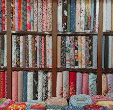 A colorful assortment of fabric rolls neatly arranged on wooden shelves. The fabrics feature various patterns and designs, including floral, geometric, and abstract motifs, with a wide range of colors from vibrant reds and blues to soft pastels. Each roll is tightly coiled, showcasing the diversity and richness of textures and styles.