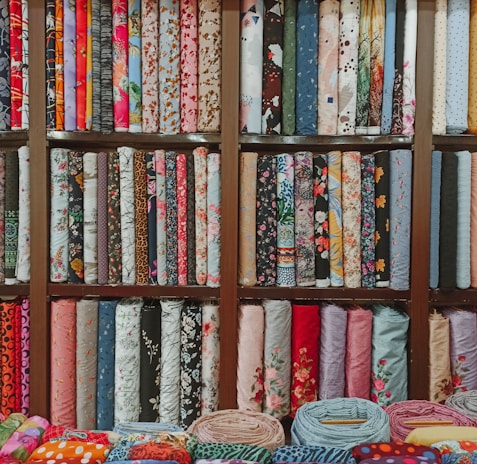 A colorful assortment of fabric rolls neatly arranged on wooden shelves. The fabrics feature various patterns and designs, including floral, geometric, and abstract motifs, with a wide range of colors from vibrant reds and blues to soft pastels. Each roll is tightly coiled, showcasing the diversity and richness of textures and styles.