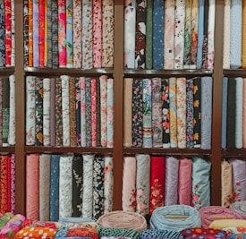 A colorful assortment of fabric rolls neatly arranged on wooden shelves. The fabrics feature various patterns and designs, including floral, geometric, and abstract motifs, with a wide range of colors from vibrant reds and blues to soft pastels. Each roll is tightly coiled, showcasing the diversity and richness of textures and styles.