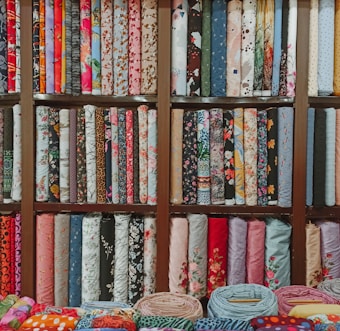 A colorful assortment of fabric rolls neatly arranged on wooden shelves. The fabrics feature various patterns and designs, including floral, geometric, and abstract motifs, with a wide range of colors from vibrant reds and blues to soft pastels. Each roll is tightly coiled, showcasing the diversity and richness of textures and styles.