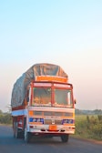 A large, colorful truck is driving on a rural road. It is heavily loaded with goods covered by a tarp. The truck displays traditional decorative elements, including bright colors and cultural motifs. It is moving along a road bordered by green fields and bushes, under a clear blue sky.