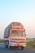 A large, colorful truck is driving on a rural road. It is heavily loaded with goods covered by a tarp. The truck displays traditional decorative elements, including bright colors and cultural motifs. It is moving along a road bordered by green fields and bushes, under a clear blue sky.