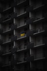 A dark, multi-story apartment building facade with repeating balconies. Each balcony has railings and a few have items like bicycles or chairs. A bright, yellow and blue flag hangs prominently on one balcony, standing out against the muted surroundings.