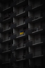 A dark, multi-story apartment building facade with repeating balconies. Each balcony has railings and a few have items like bicycles or chairs. A bright, yellow and blue flag hangs prominently on one balcony, standing out against the muted surroundings.