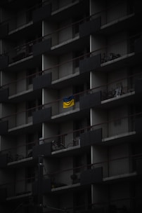 A dark, multi-story apartment building facade with repeating balconies. Each balcony has railings and a few have items like bicycles or chairs. A bright, yellow and blue flag hangs prominently on one balcony, standing out against the muted surroundings.