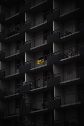 A dark, multi-story apartment building facade with repeating balconies. Each balcony has railings and a few have items like bicycles or chairs. A bright, yellow and blue flag hangs prominently on one balcony, standing out against the muted surroundings.