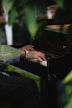 A person is playing an old black piano surrounded by lush green foliage. The person has a wristband with the text 'GIVE' on it and is wearing a colorful patterned shirt.