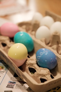 A carton holds a mix of colored and white eggs. The colored eggs display pastel shades of pink, yellow, blue, and turquoise, with one decorated with a zigzag pattern. The carton rests on newspaper sheets, partially visible at the edges.