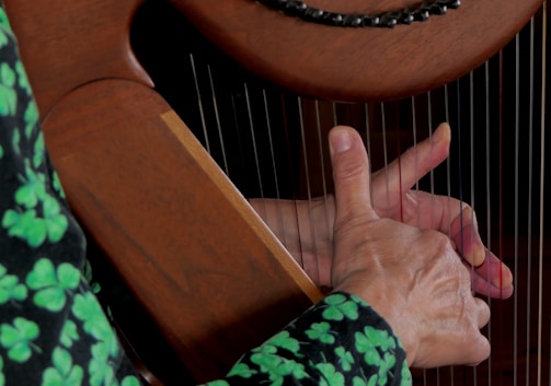 A person's hand is placed on the strings of a wooden harp, with fingers poised to pluck or strum. The natural grain of the wood and the delicate strings are visible, highlighting the craftsmanship of the instrument. The person is wearing a dark garment with bright green clover patterns.