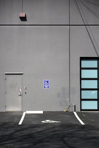 A gray building exterior with a metal door and a blue handicapped parking sign. The ground features a marked parking space indicating accessibility, while the wall shows some electrical wires and plant growth at the base.