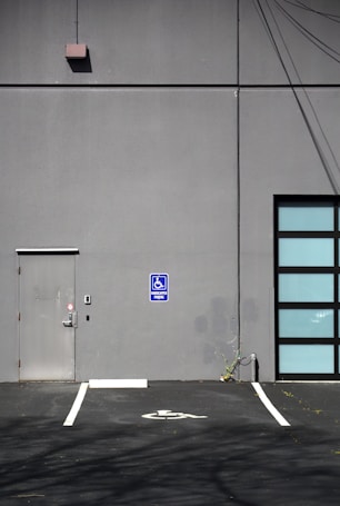 A gray building exterior with a metal door and a blue handicapped parking sign. The ground features a marked parking space indicating accessibility, while the wall shows some electrical wires and plant growth at the base.
