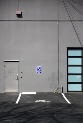 A gray building exterior with a metal door and a blue handicapped parking sign. The ground features a marked parking space indicating accessibility, while the wall shows some electrical wires and plant growth at the base.