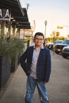 A young boy stands confidently on a sidewalk in an urban setting, wearing a plaid blazer, patterned shirt, and ripped jeans. He is positioned near a row of plants, with parked cars and a building in the background.