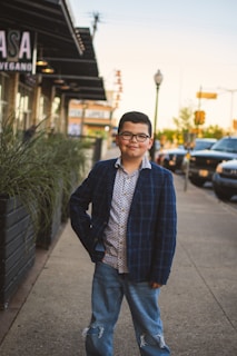 A young boy stands confidently on a sidewalk in an urban setting, wearing a plaid blazer, patterned shirt, and ripped jeans. He is positioned near a row of plants, with parked cars and a building in the background.