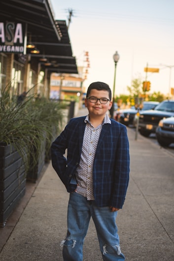 A young boy stands confidently on a sidewalk in an urban setting, wearing a plaid blazer, patterned shirt, and ripped jeans. He is positioned near a row of plants, with parked cars and a building in the background.