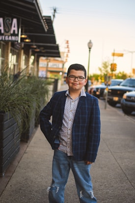 A young boy stands confidently on a sidewalk in an urban setting, wearing a plaid blazer, patterned shirt, and ripped jeans. He is positioned near a row of plants, with parked cars and a building in the background.