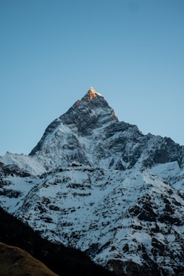 A towering snow-capped mountain peak with a clear sky background. The peak catches the golden light at the very top, suggesting the warm glow of sunrise or sunset. The rugged terrain and snowy slopes are clearly visible, creating a serene and majestic landscape.