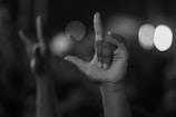 A hand in focus is positioned with fingers forming the sign for 'I love you' in American Sign Language, against a blurry background with scattered light spots.