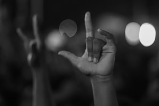 A hand in focus is positioned with fingers forming the sign for 'I love you' in American Sign Language, against a blurry background with scattered light spots.