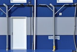 A section of a blue and gray building features a white door centered between two rows of windows. Gray pipes run vertically and horizontally across the facade, creating a geometric pattern. A sign with text is mounted near the bottom right corner.