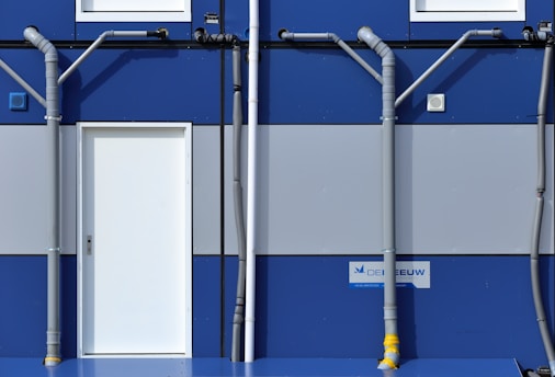 A section of a blue and gray building features a white door centered between two rows of windows. Gray pipes run vertically and horizontally across the facade, creating a geometric pattern. A sign with text is mounted near the bottom right corner.
