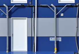 A section of a blue and gray building features a white door centered between two rows of windows. Gray pipes run vertically and horizontally across the facade, creating a geometric pattern. A sign with text is mounted near the bottom right corner.