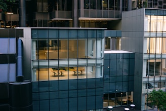 A modern office building facade with large glass windows showing interior spaces. The reflection and transparency reveal office desks and chairs inside. The architecture is characterized by a mix of steel and glass panels, creating a sleek and contemporary look.