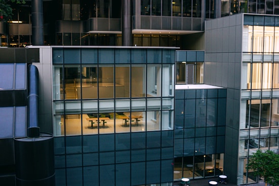 A modern office building facade with large glass windows showing interior spaces. The reflection and transparency reveal office desks and chairs inside. The architecture is characterized by a mix of steel and glass panels, creating a sleek and contemporary look.