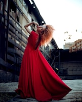 A model with vibrant red hair poses dramatically in a long, flowing red dress. The setting appears to be an urban environment with industrial building elements in the background. Her stance is dynamic and fashion-forward.