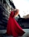 A model with vibrant red hair poses dramatically in a long, flowing red dress. The setting appears to be an urban environment with industrial building elements in the background. Her stance is dynamic and fashion-forward.