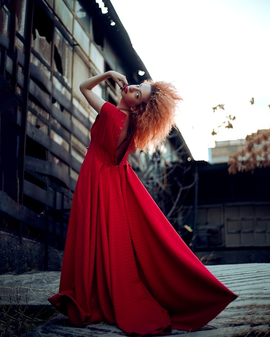 A model with vibrant red hair poses dramatically in a long, flowing red dress. The setting appears to be an urban environment with industrial building elements in the background. Her stance is dynamic and fashion-forward.