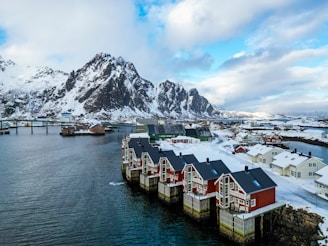 Red wooden houses with black roofs are positioned along a snowy shoreline, adjacent to a calm, blue body of water. Snow-capped mountains rise prominently in the background under a partially cloudy sky, creating a serene and picturesque winter landscape.