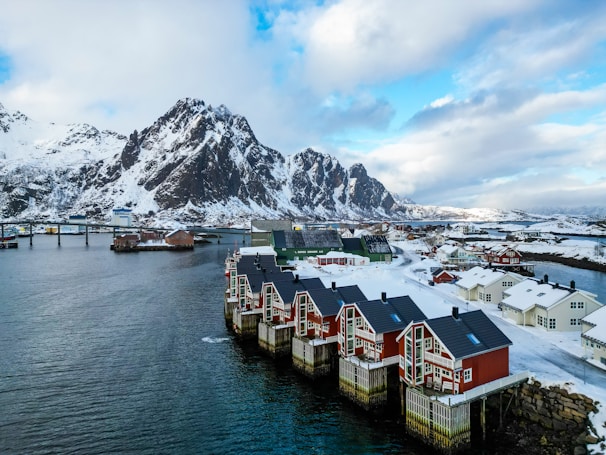 Red wooden houses with black roofs are positioned along a snowy shoreline, adjacent to a calm, blue body of water. Snow-capped mountains rise prominently in the background under a partially cloudy sky, creating a serene and picturesque winter landscape.