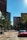 A city street with tall buildings lining both sides, including one with the signage 'K&L Gates'. Trees are present along the sidewalk, and several vehicles, including a red car, are on the road. The scene appears to be taken during daylight under a clear blue sky.