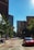A city street with tall buildings lining both sides, including one with the signage 'K&L Gates'. Trees are present along the sidewalk, and several vehicles, including a red car, are on the road. The scene appears to be taken during daylight under a clear blue sky.