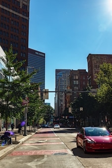 A city street with tall buildings lining both sides, including one with the signage 'K&L Gates'. Trees are present along the sidewalk, and several vehicles, including a red car, are on the road. The scene appears to be taken during daylight under a clear blue sky.