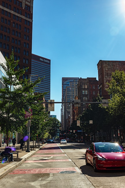 A city street with tall buildings lining both sides, including one with the signage 'K&L Gates'. Trees are present along the sidewalk, and several vehicles, including a red car, are on the road. The scene appears to be taken during daylight under a clear blue sky.