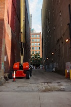 An orange construction lift is parked in a narrow alley between tall brick and concrete buildings. The alley is paved with a cracked surface and has orange traffic cones around the lift. In the distance, a building with a modern glass facade is visible, along with some trees and a few people walking.