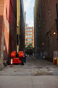 An orange construction lift is parked in a narrow alley between tall brick and concrete buildings. The alley is paved with a cracked surface and has orange traffic cones around the lift. In the distance, a building with a modern glass facade is visible, along with some trees and a few people walking.