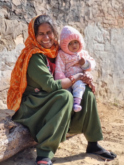 A joyful woman wearing an orange scarf and a green dress sits on a log holding a warmly dressed baby in a pink hooded outfit. Both are seated against a weathered stone wall on sunlit, sandy ground.
