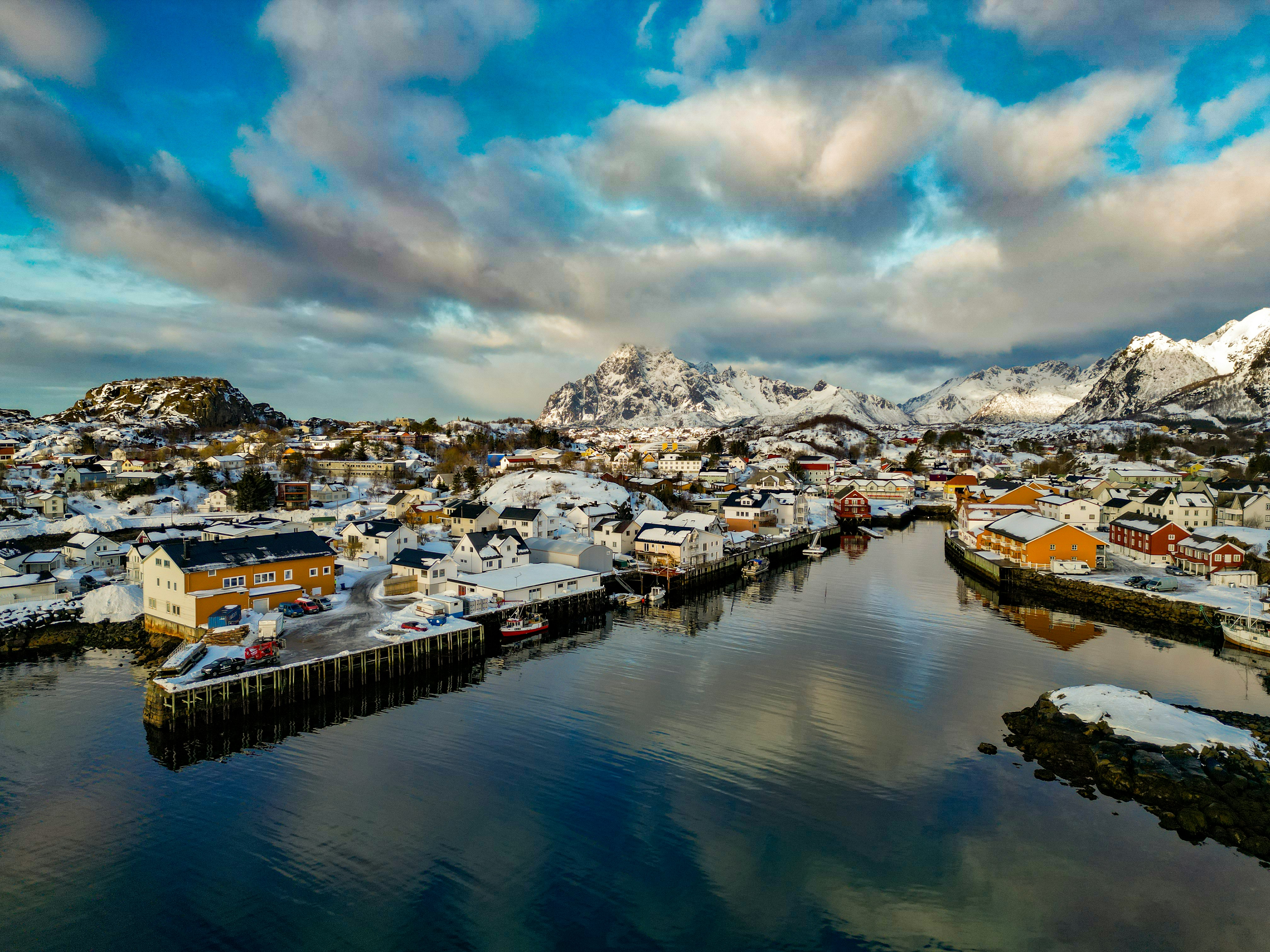 an aerial view of a small town with mountains in the background, A little harbor in Lofoten Norway. Photo taken with drone in good sunlight. Snow-covered mountains in the background. Rorbuer