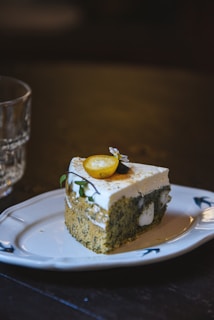 A slice of cake featuring a dark speckled sponge base topped with a creamy frosting is placed on a decorative white plate. The cake is garnished with a small piece of yellow fruit and edible flowers. In the background, a clear glass with textured patterns is visible, sitting on a dark wooden surface.