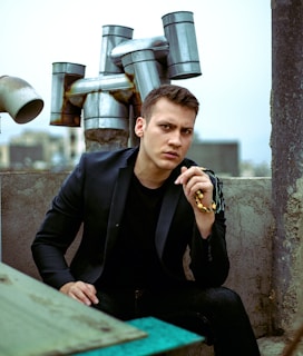 A young man with short hair is sitting with a pensive expression on his face, dressed in a black jacket and shirt. He is holding a beaded rosary in one hand. The background features an industrial setting with multiple exhaust pipes protruding from a structure and a blurred urban skyline in the distance.
