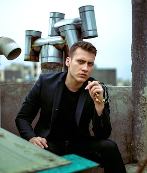 A young man with short hair is sitting with a pensive expression on his face, dressed in a black jacket and shirt. He is holding a beaded rosary in one hand. The background features an industrial setting with multiple exhaust pipes protruding from a structure and a blurred urban skyline in the distance.