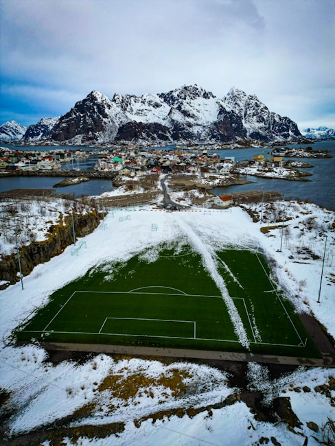A green soccer field is partly covered with snow, surrounded by a snowy landscape. In the background, a scenic view of mountains with snow-covered peaks is visible, along with a small coastal village with colorful houses and patches of open water.